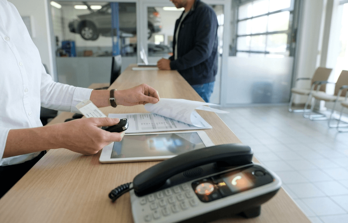 Mitarbeiterin am Servicetresen mit Autoschlüssel und Auftragsblatt neben Tablet und Telefon, dahinter ein Kunde